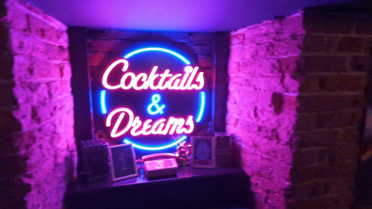 Różowo-niebieski neon z napisem Cocktails and Dreams na ceglanej ścianie, pod nim retro telefon i ramki ze zdjęciami
