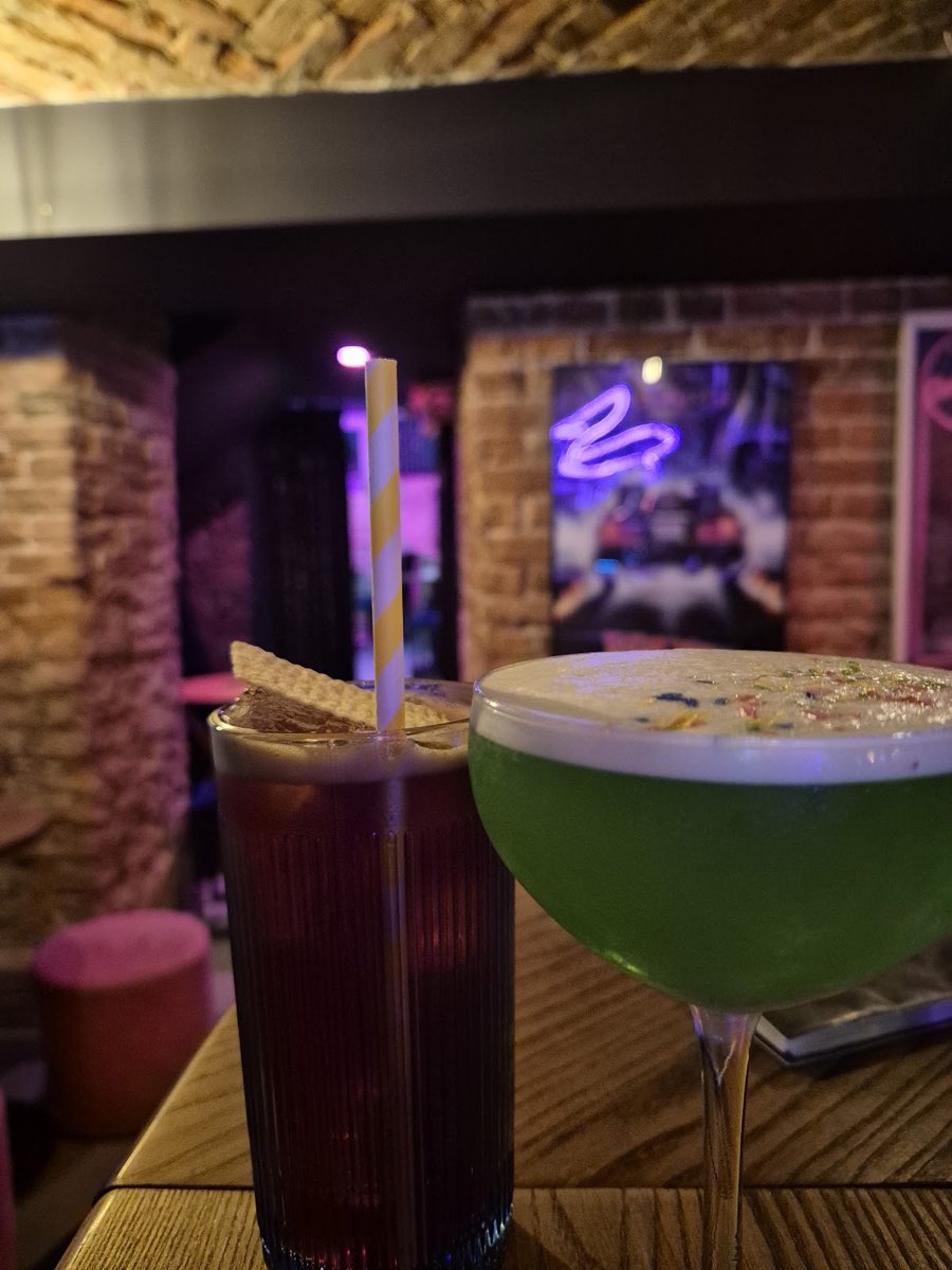 Zielony koktajl w kieliszku coupe i czerwony drink w wysokiej szklance na drewnianym barze, w tle plakat i ceglana ściana
